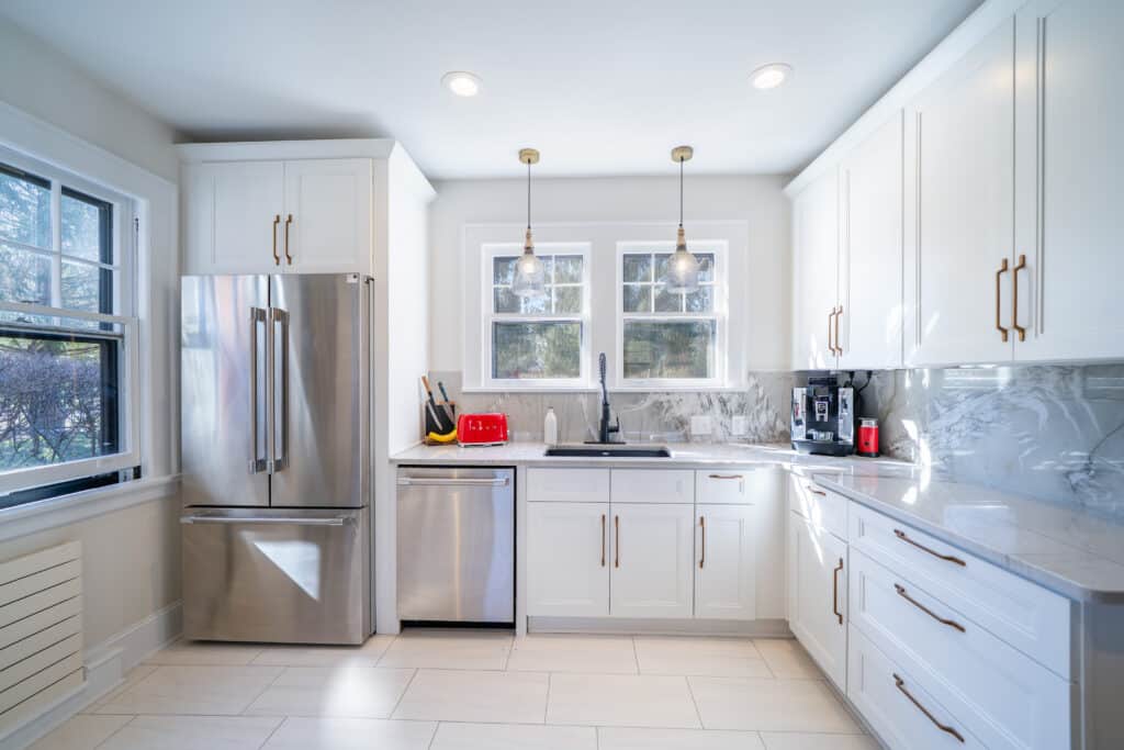Bright modern kitchen with white cabinets, stainless steel appliances, marble backsplash, pendant lights, and sunlight coming through two windows above the sink.